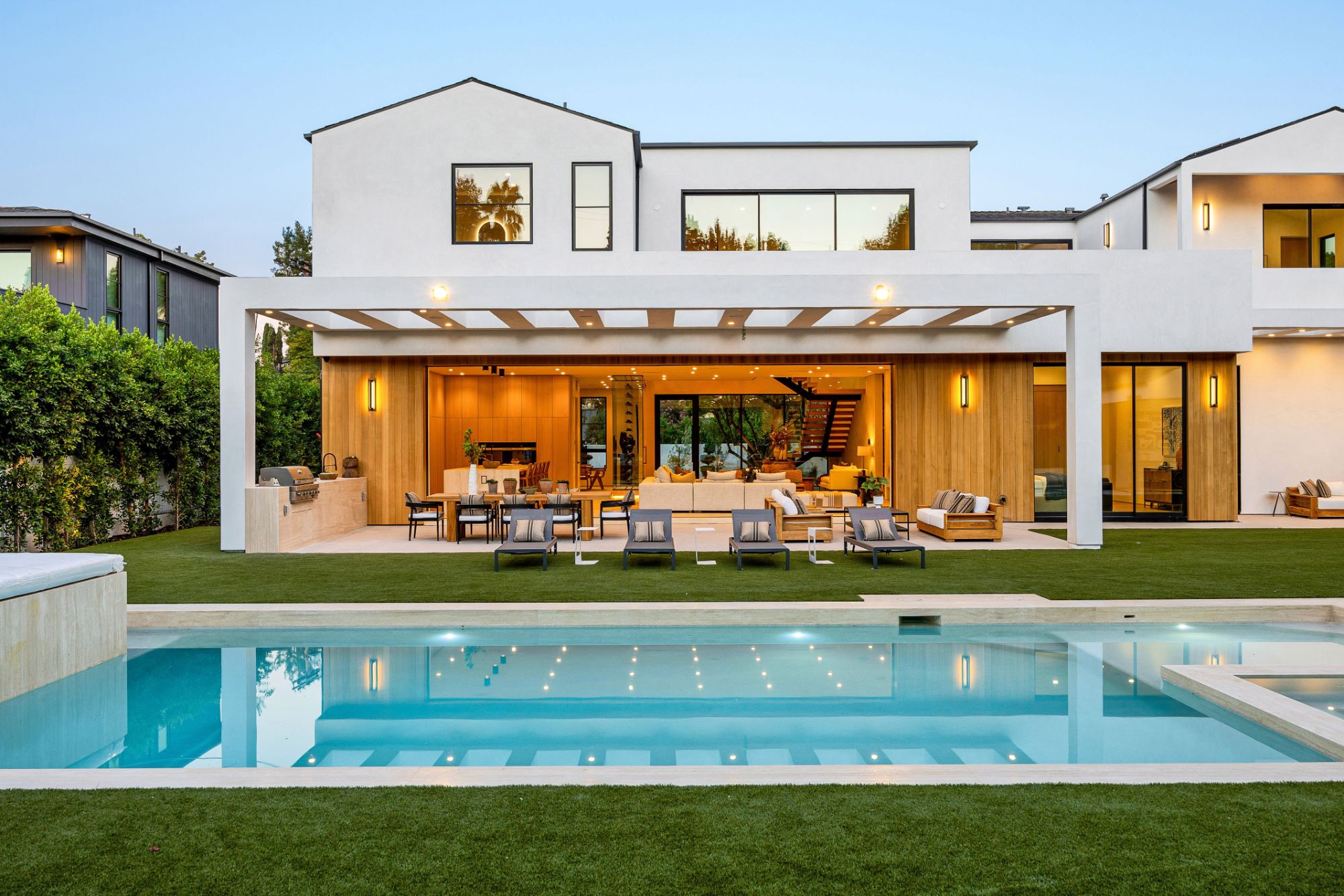 White modern home with a covered terrace and pool at dusk, its interior lights on